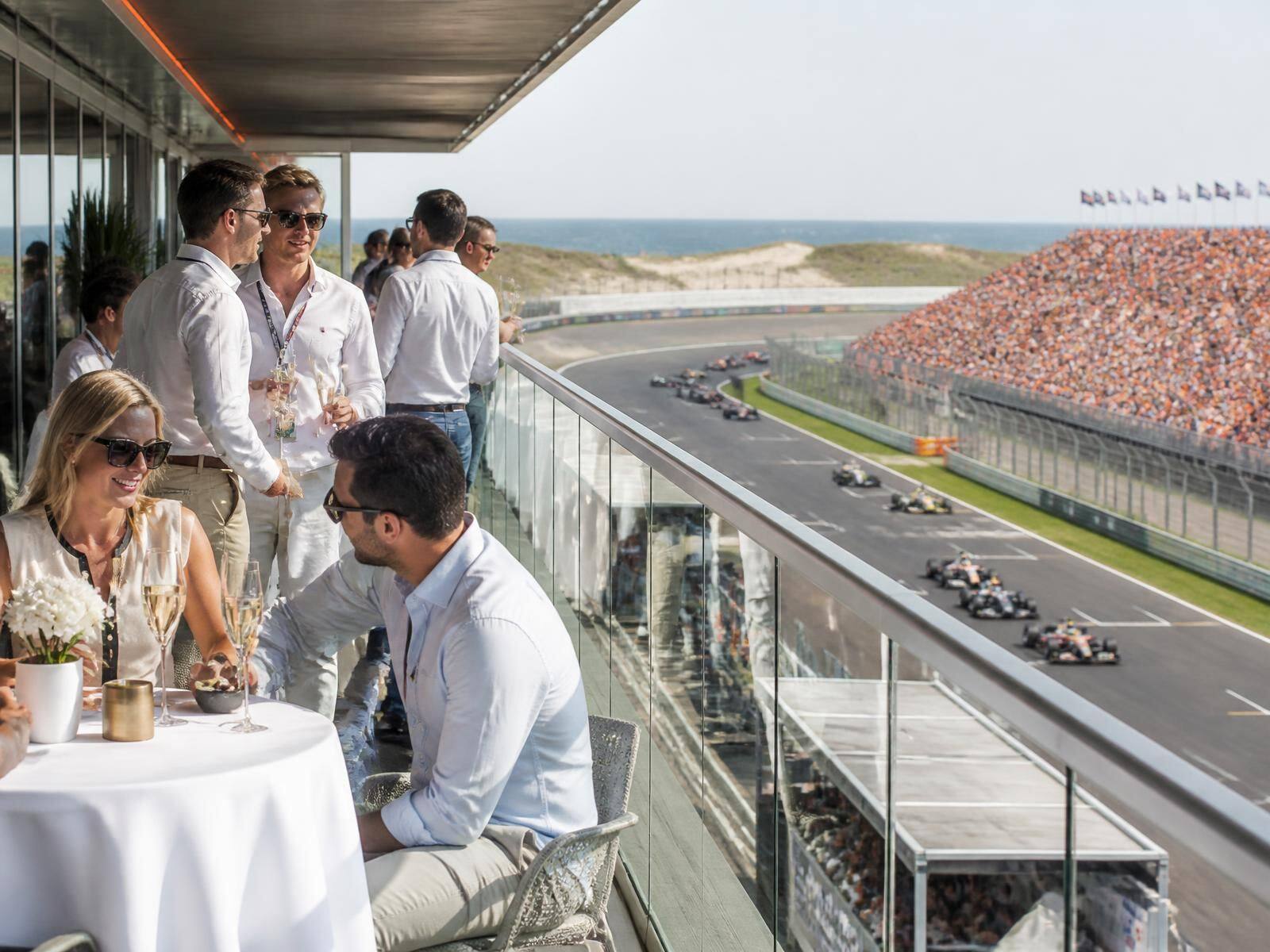 Gasten op het Paddock Club terras boven de pitboxen met de raceauto's op de baan